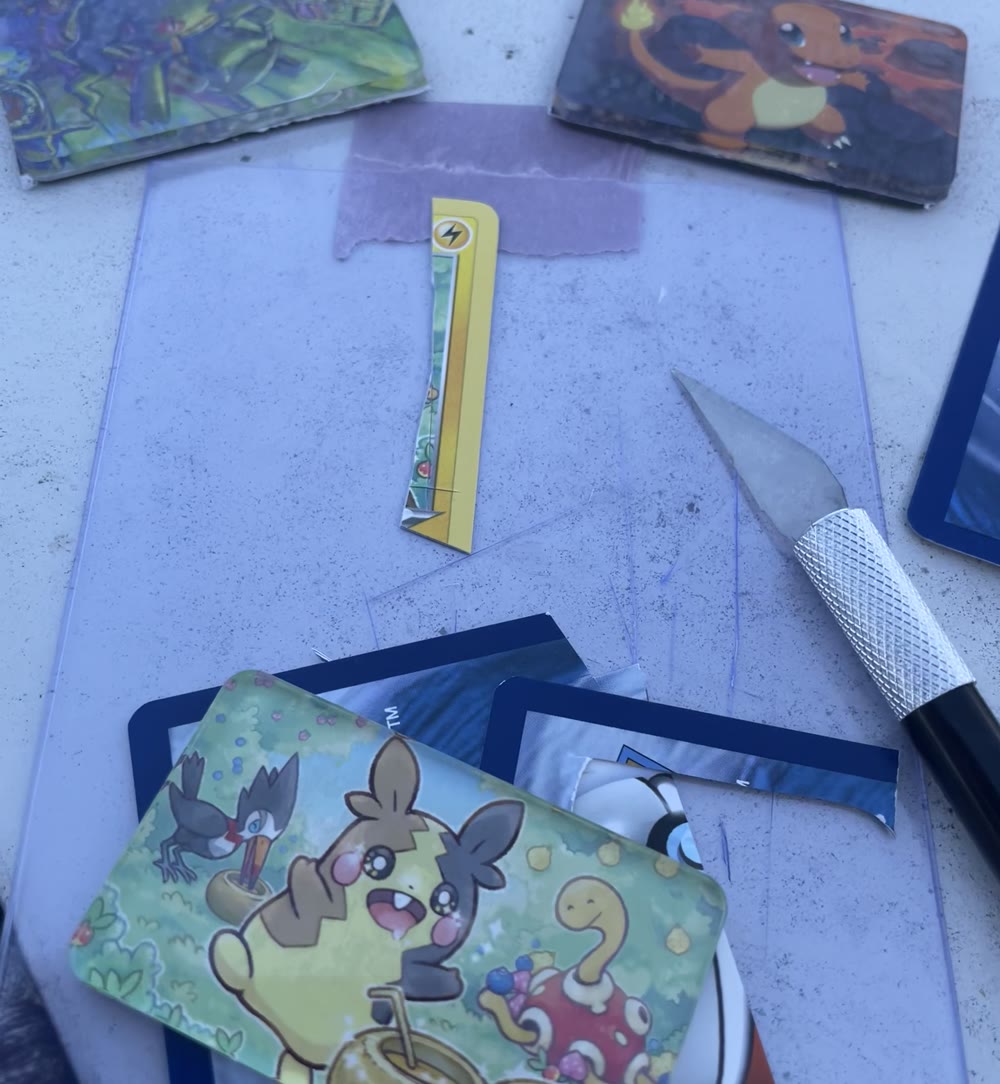 A work surface with a clear acrylic sheet, a craft knife, trimmed offcuts of Pokémon cards, and two finished acrylic magnets, one showing Charmander and one a busy green jungle scene