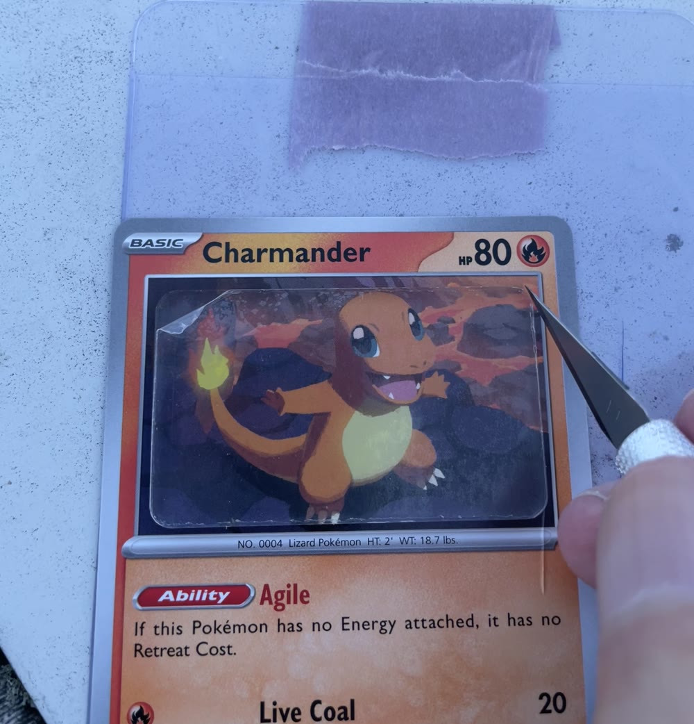 A Charmander card lying on a cutting surface with a craft knife blade resting along the edge of its artwork window, before any cut is made