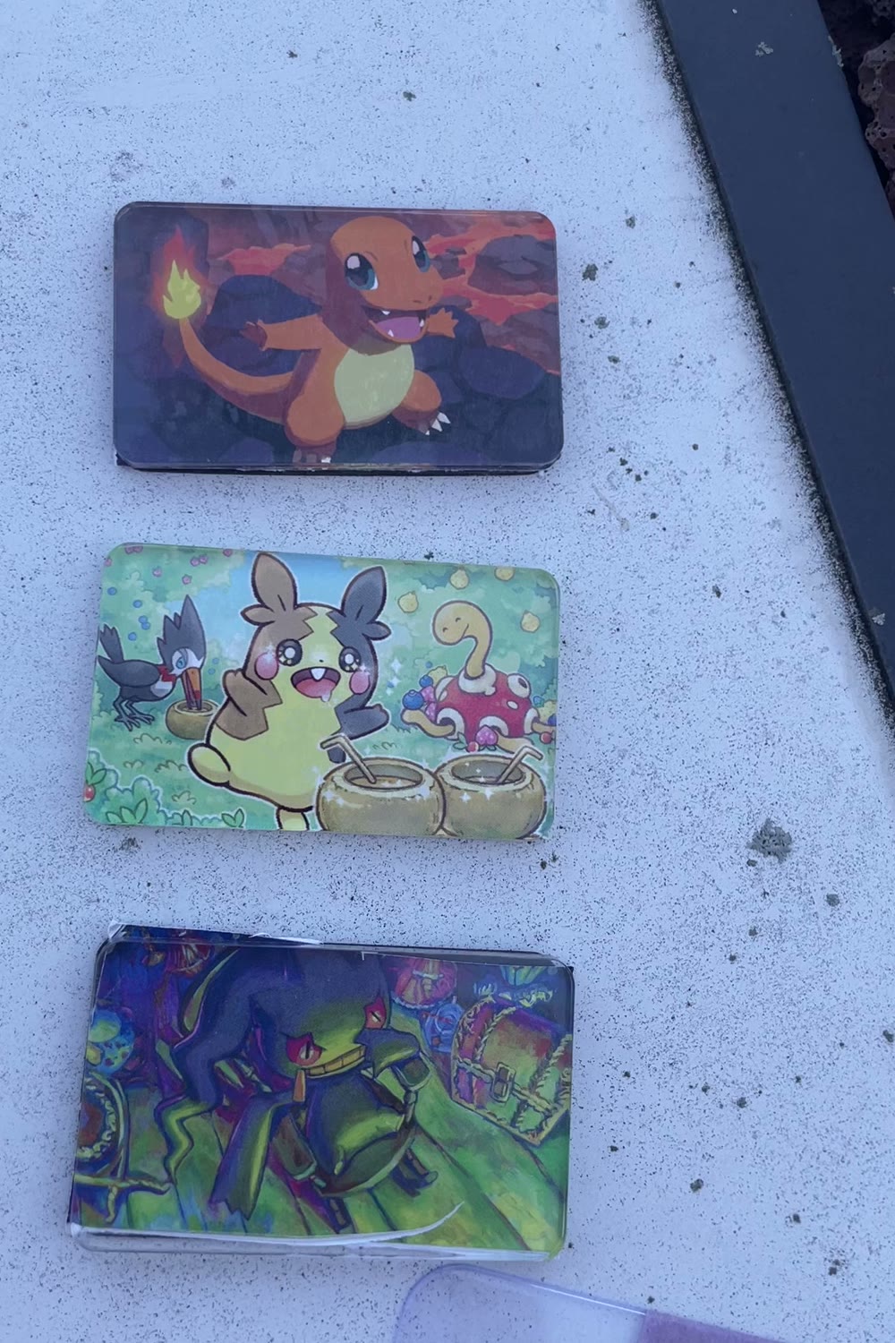 Three finished Pokémon card magnets laid out on a light table: Charmander on lava rock, Morpeko at a drinks stand, and a Banette treasure-room scene whose trimmed edge is visibly ragged